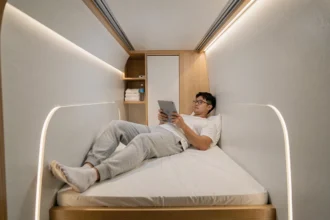 Intérieur d'une capsule d'hôtel japonais avec un homme allongé lisant une tablette dans un espace compact de 2 mètres sur 1 mètre